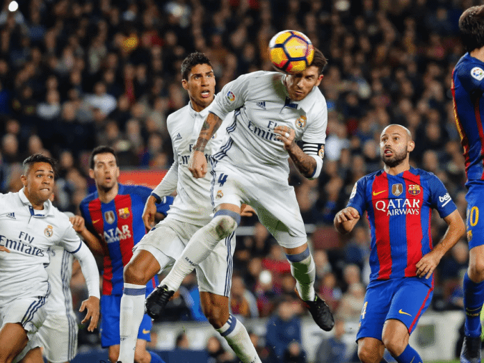 A tense El Clásico moment where Real Madrid's Sergio Ramos wins an aerial duel with Barcelona defenders